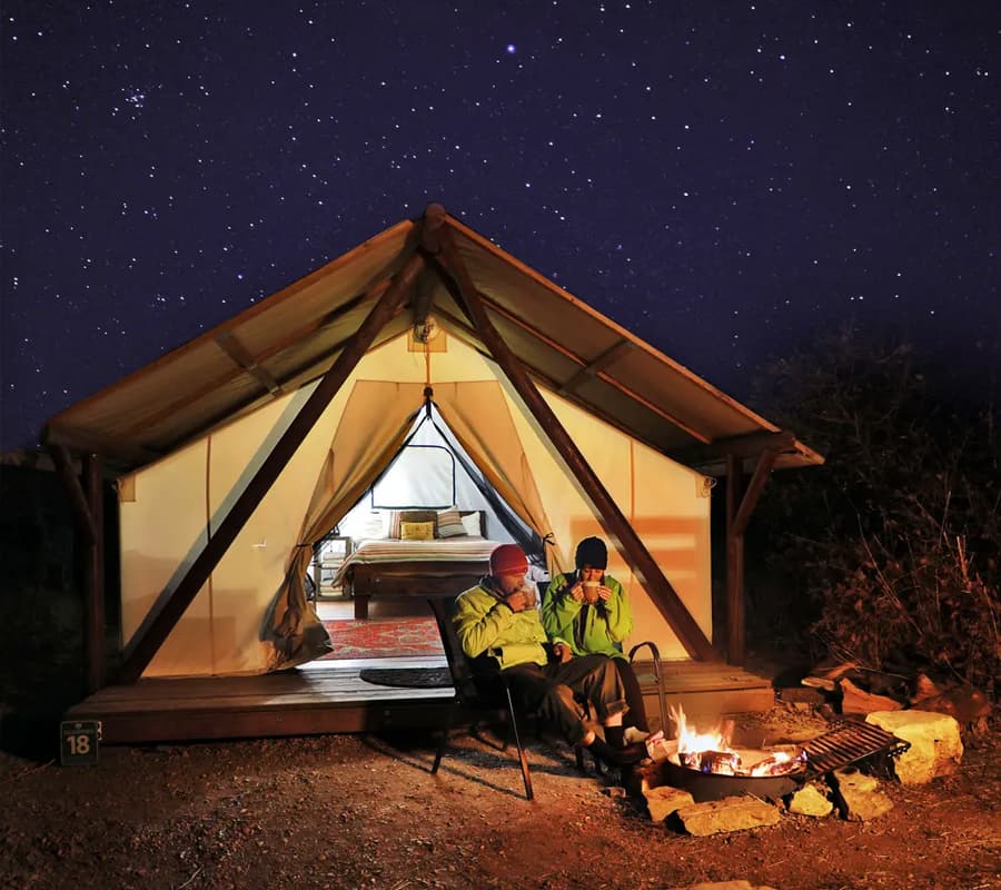 Two people sit by a campfire in front of a glamping tent under a starry night sky.