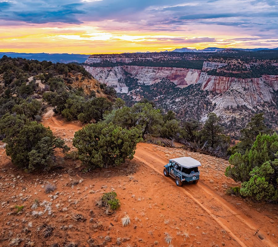 A vehicle drives along a winding dirt road amid scenic cliffs and a vibrant sunset.