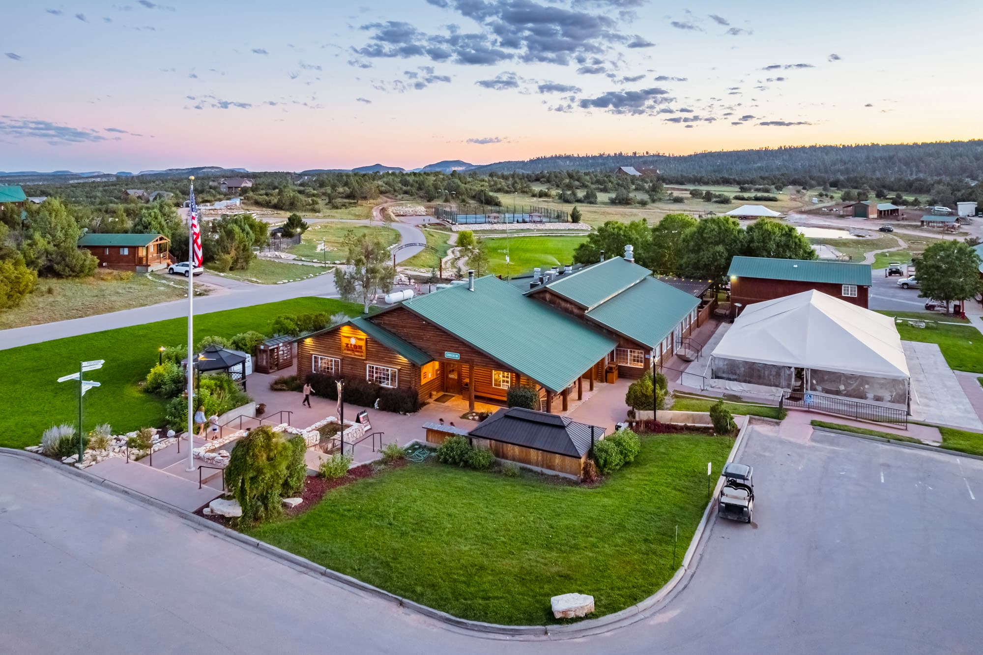 Aerial view of a rustic lodge with green roofs surrounded by grass and trees at sunset.